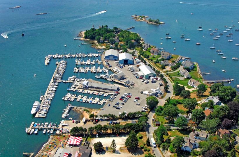 An aerial view of a marina with numerous boats, docks, adjacent buildings, and greenery, surrounded by a large body of water.